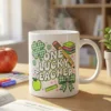 Cozy St Patrick's Day teacher coffee mug mockup with steaming hot beverage, featuring retro coquette "One Lucky Teacher" design with gingham shamrock and bow on wooden desk with lesson planner