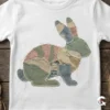 White toddler t-shirt featuring a camouflage Easter bunny design on a rustic wood background.