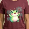 Maroon adult t-shirt mockup displaying a happy green frog celebrating with colorful fireworks in the background.