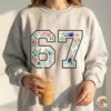Woman wearing an ash grey sweatshirt featuring the viral floral 67 number design, holding an iced coffee in a sunny setting.