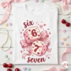 Flat lay photography of a white Valentine's Day t-shirt with the Six Seven dice design, surrounded by pink satin ribbons, pearls, and cherries.