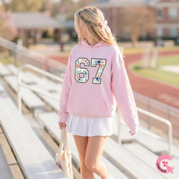 Girl with a bow in her hair wearing a light pink hoodie with the floral 67 design standing on school stadium bleachers.