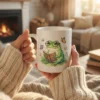 Cozy lifestyle mockup of a person in a knit sweater holding a white ceramic coffee mug printed with a cute watercolor frog reading a book.