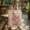 A canvas tote bag hanging on a wooden chair outdoors, featuring the Six Seven dice and bow design, with a water bottle and notebook inside.