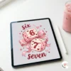 An iPad Pro lying on a white desk displaying the Six Seven dice and bow digital art, next to an Apple Pencil and pink iced coffee.