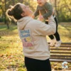 Mother laughing and lifting toddler in park wearing a beige oversized hoodie with String Cheese Mama Era sublimation design.