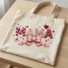 Natural heavy canvas tote bag printed with the 3D textured Mama pink bow design, lying flat on a wooden desk.