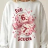 A blonde model wearing a white crewneck sweatshirt featuring the viral Six Seven 67 dice design with pink coquette bows and hearts.