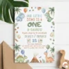 Flat lay of watercolor One-a-Saurus 1st birthday invitation with wooden dino toy and kraft envelope on white wood table.